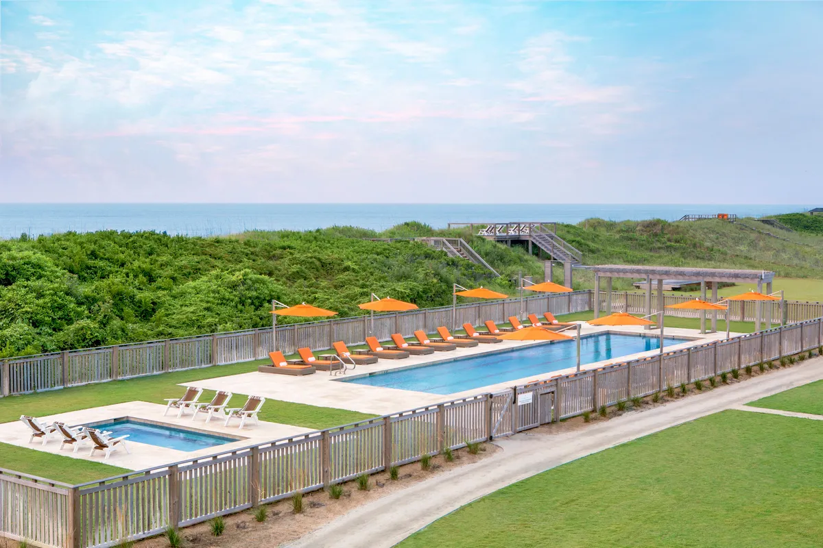 A sun-drenched swimming pool with plush lounge chairs and signature orange umbrellas, overlooking the ocean. The space is set up for morning yoga sessions, sunbathing, swimming, and leisurely gatherings, all with a view of the horizon.