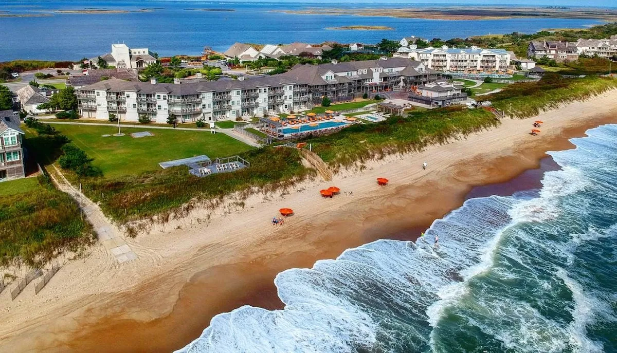 A tranquil exterior scene featuring the Sanderling Resort's beachfront with inviting hotel architecture, a swimming pool lined with umbrellas, and the glistening ocean as a backdrop. Guests are lounging beside the pool, sunbathing, swimming, and strolling along the sandy shore.