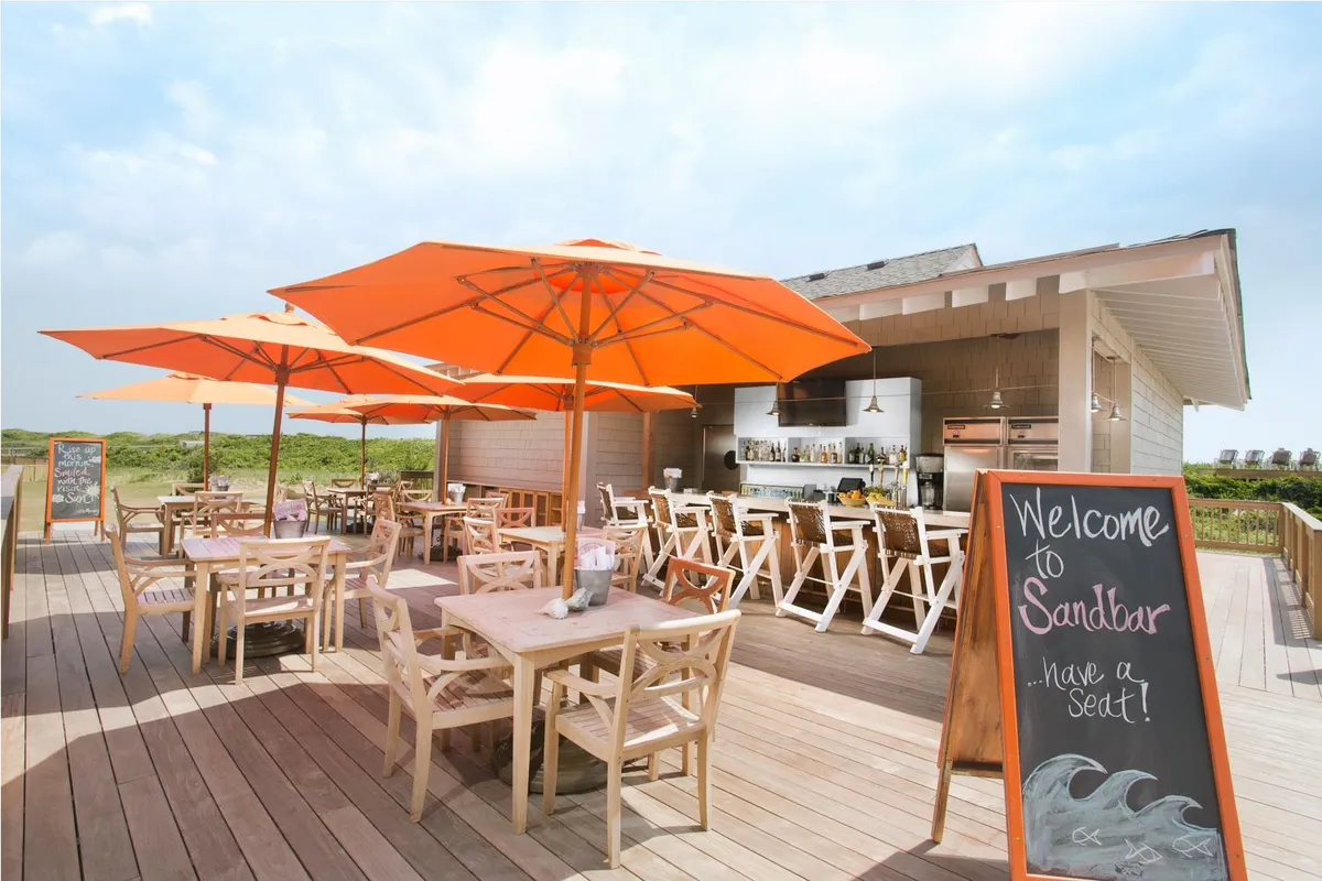 An inviting outdoor dining area with orange umbrellas shading wooden tables, adjacent to a lively bar. The setting is designed for socializing, al fresco dining, and enjoying fresh seafood, all with the soft glow of sunset in the background.