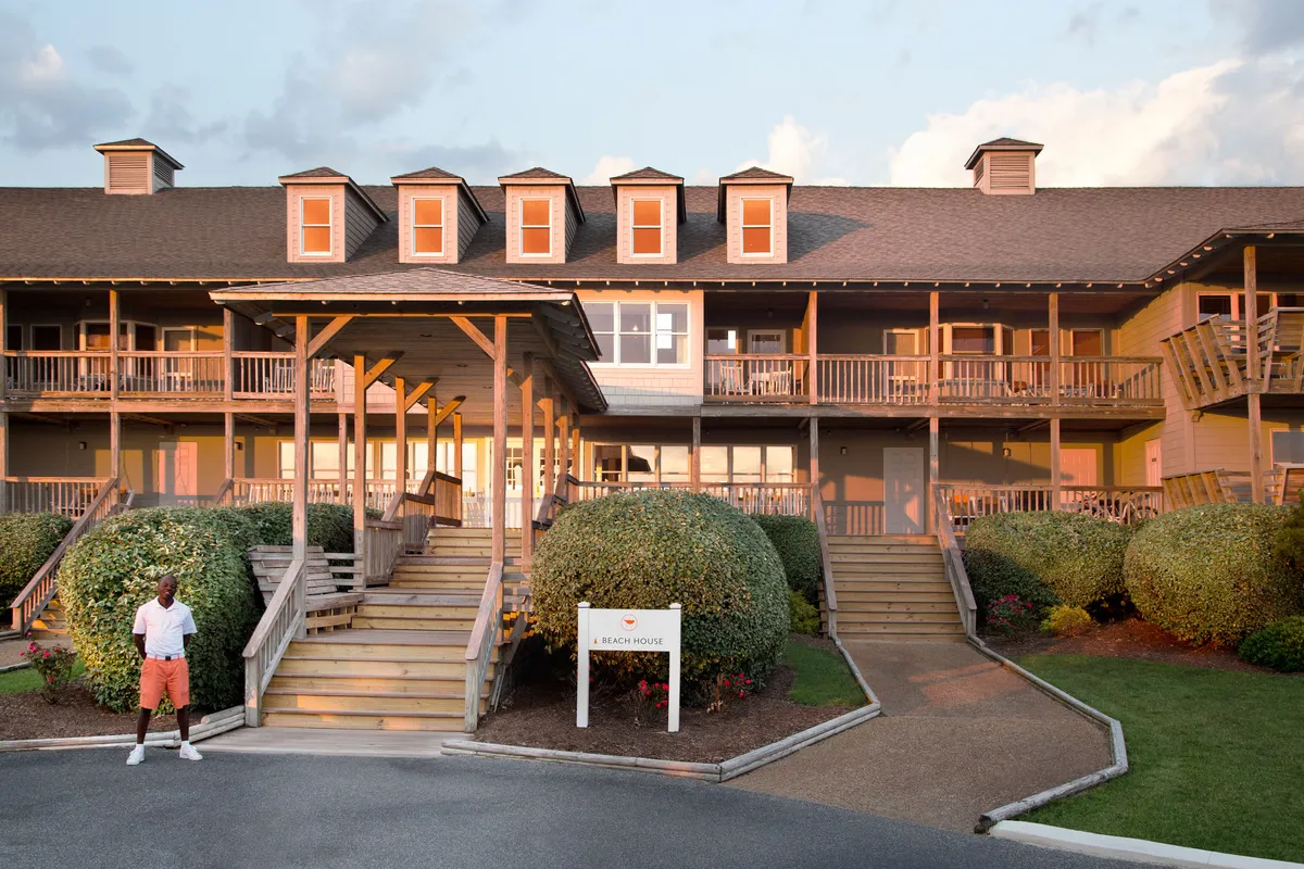 This image displays the inviting exterior of the property, featuring a welcoming building facade, well-maintained entrance steps, landscaped bushes, and visible signage. The scene is bathed in the warm light of sunset, suggesting a tranquil atmosphere ideal for relaxation and social gatherings. The porch and open yard provide perfect spaces for group activities such as shared meals, evening games, or enjoying cocktails at sunset, all set against the backdrop of natural beauty.
