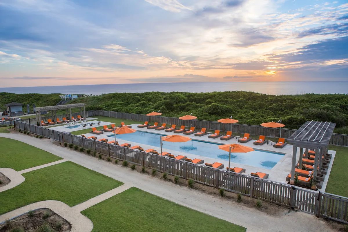A sparkling outdoor swimming pool surrounded by vibrant orange umbrellas, with lounge chairs arranged for relaxing and socializing. In the distance, the sun sets over the ocean, creating a dreamy and carefree ambiance. Some guests are swimming or unwinding poolside, suggesting communal fun and relaxation.