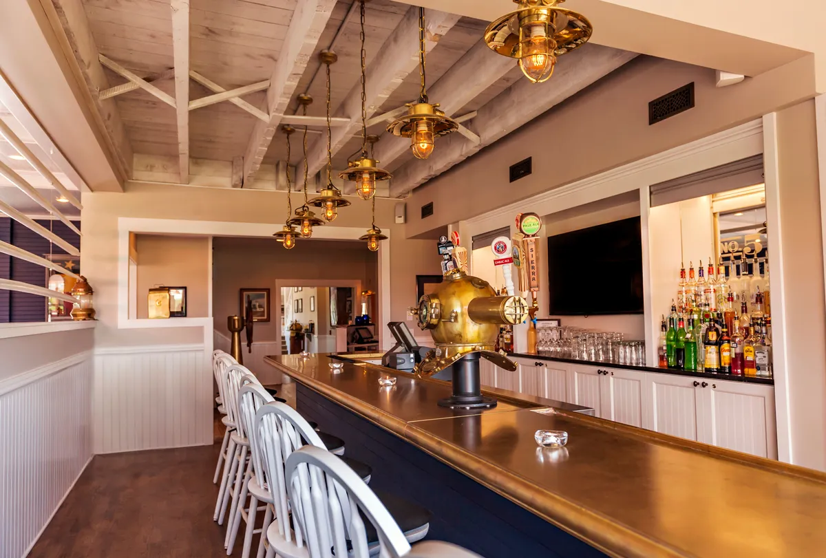 A stylish bar area with a long counter, hanging pendant lights, decorative wall art, and an array of bottled drinks on display. The atmosphere is inviting and social, ideal for relaxed gatherings or evening celebrations.