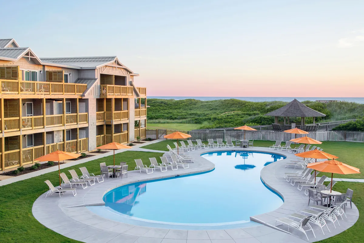 A vibrant resort pool area featuring sparkling water, cushioned lounge chairs, orange umbrellas for shade, and a beautifully landscaped lawn, with the resort building in the background. The atmosphere is lively and inviting, ideal for both relaxation and group gatherings.