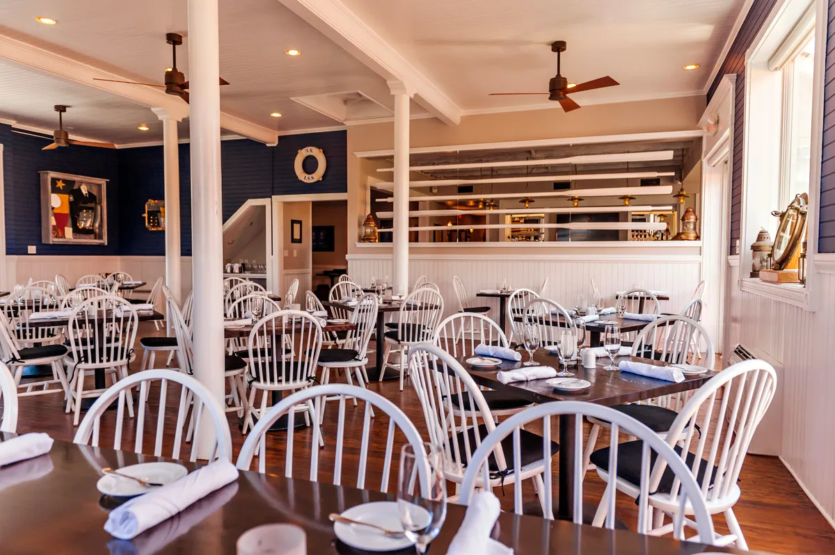 A cozy and inviting dining space with white wooden chairs, dark wooden tables, blue accent walls adorned with artwork, and brass fixtures at the bar. The scene radiates warmth and encourages lingering conversations over shared meals.