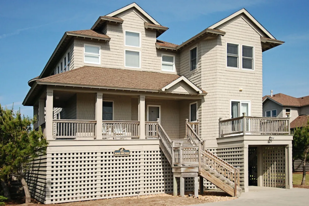 The image showcases the inviting front porch of a beautifully designed beach house in Duck, North Carolina. Wooden steps lead up to the spacious porch, perfect for relaxing, socializing, or enjoying a sunset. The unique architectural details of the home are visible, creating a welcoming and tranquil atmosphere ideal for family gatherings and outdoor fun.