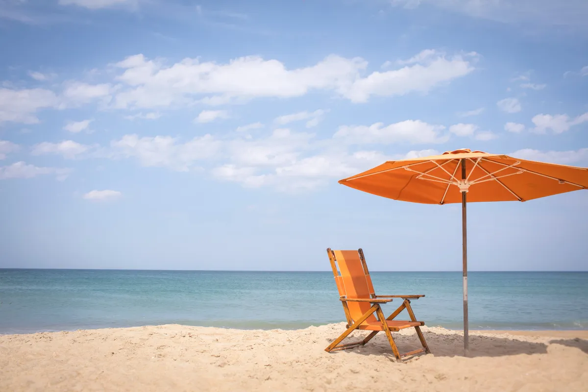 A tranquil stretch of sandy beach with a classic beach chair and umbrella set up, looking out over the gentle surf and open ocean under a sunlit sky. The scene invites relaxation and fun, with ample space for families to gather, play, and enjoy the water.