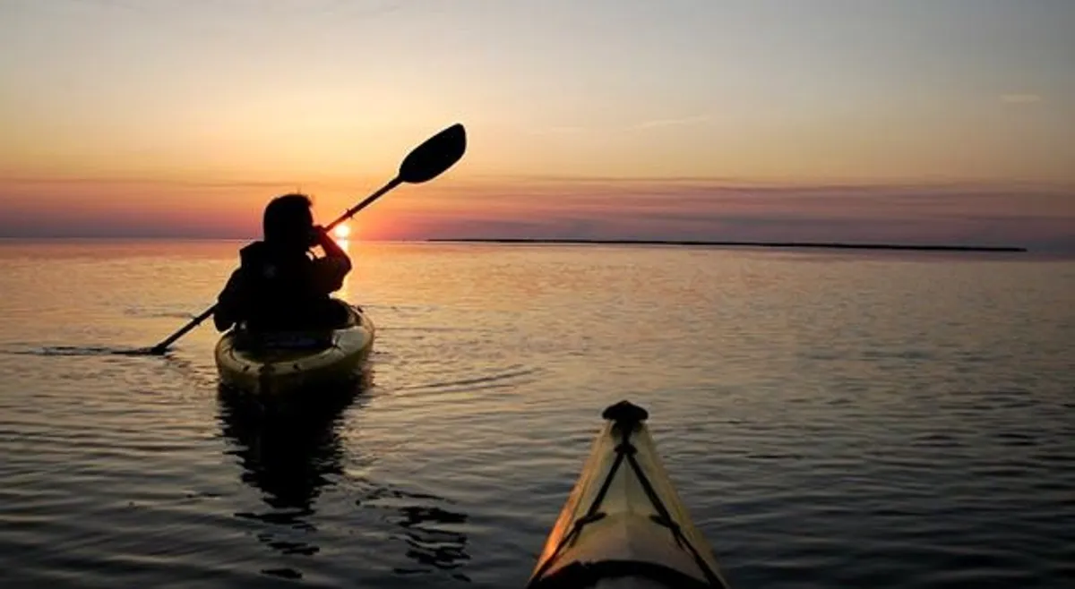 A kayaker glides across calm waters at sunset, surrounded by the expansive beauty of Currituck Sound. The warm hues and mirror-like surface evoke a sense of adventure and tranquility, inviting viewers to imagine themselves exploring the natural landscape.
