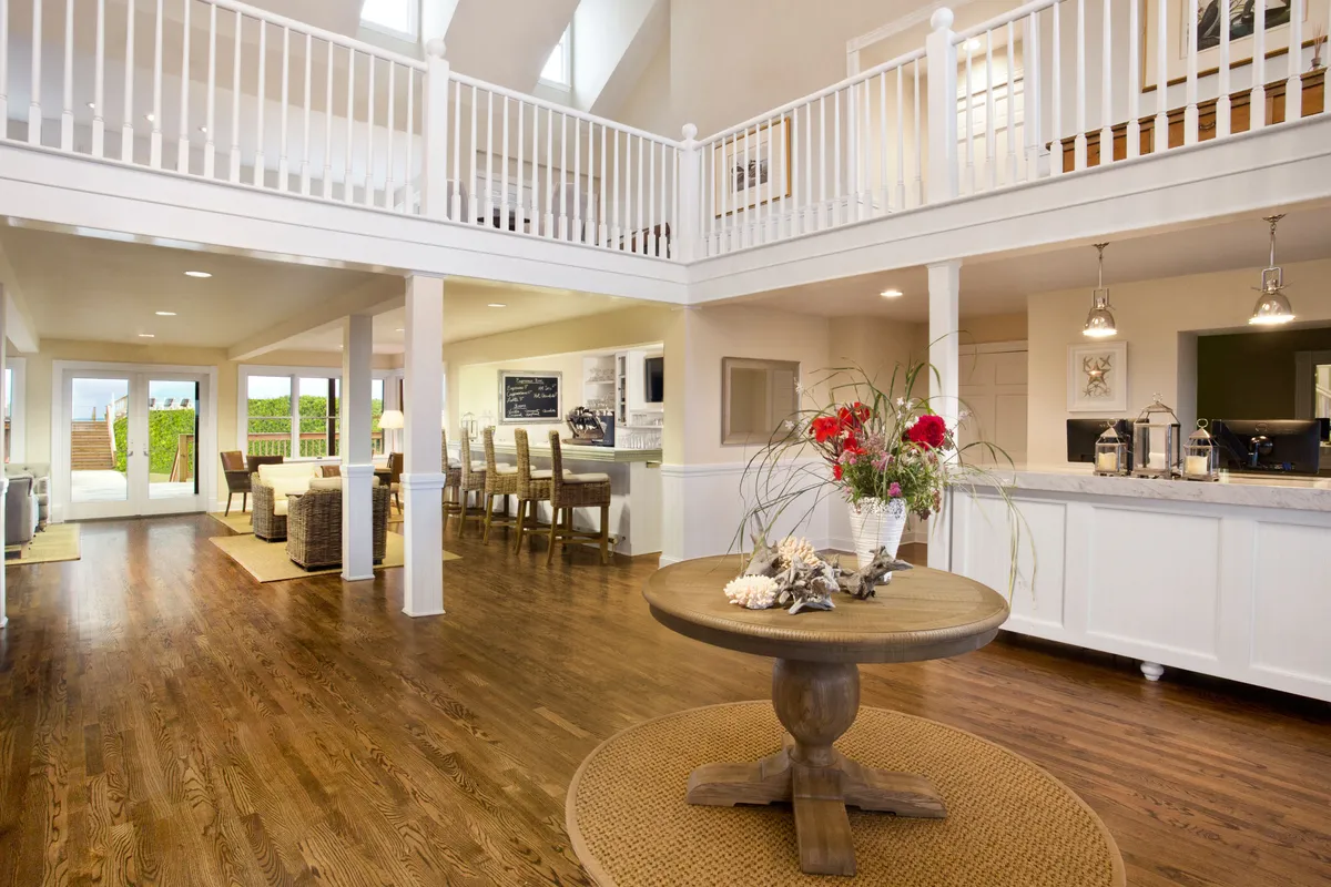 An open, airy interior with a round table adorned with a floral arrangement, adjacent to a modern kitchen. Large windows open out to an outdoor area, inviting both natural light and easy access between inside and outside. The setting encourages group meals and casual get-togethers.