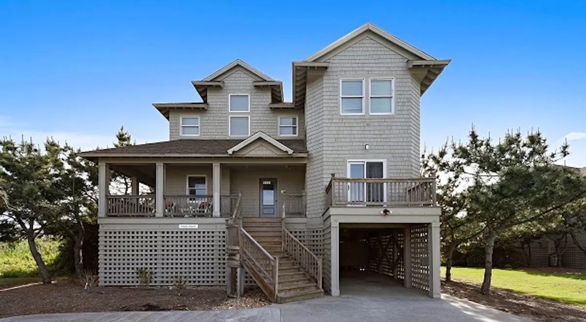 A welcoming front porch framed by lush trees, featuring wide steps, large windows, and comfortable seating. The porch invites relaxation with views of the surrounding nature and is a natural extension of the living spaces within.