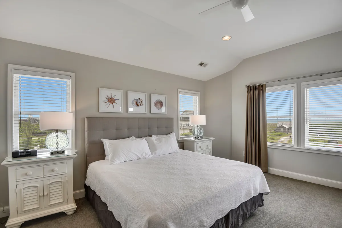 A thoughtfully styled bedroom featuring a plush bed, decorative lamps, and windows that let in ample daylight. The inviting textures and calming palette offer a perfect spot for restful sleep or quiet moments with a book.