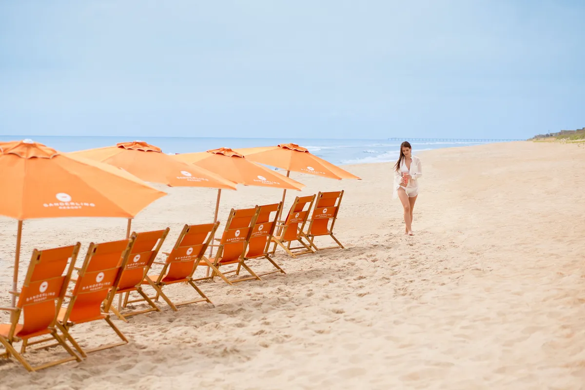 A serene private beachfront with beach chairs and umbrellas set up for relaxation, a person strolling along the shore, and gentle waves lapping at the sand. The mood is peaceful and exclusive, emphasizing privacy and connection to nature.