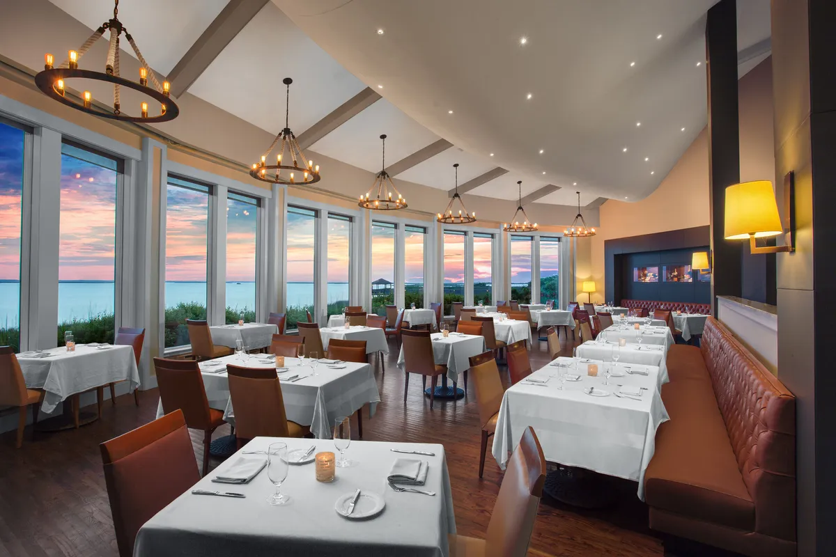 An elegant dining room with large windows offering a stunning sunset view over the water. The room features chandeliers, beautifully set tables, and a warm, welcoming ambiance that invites guests to enjoy memorable meals together.