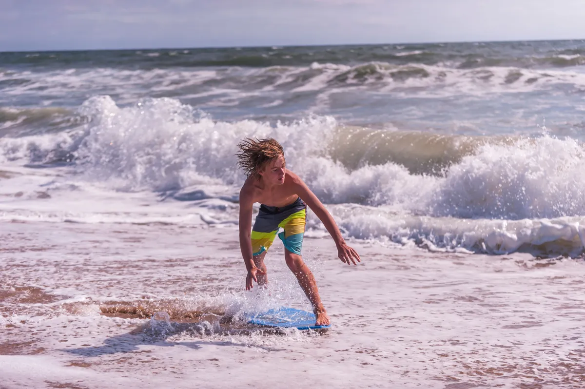 A joyful scene of a young boy riding a boogie board as waves break around him, capturing a lively moment of play in the surf. The ocean gleams in the sunlight, and the shoreline in the background hints at families and children enjoying active beach adventures.