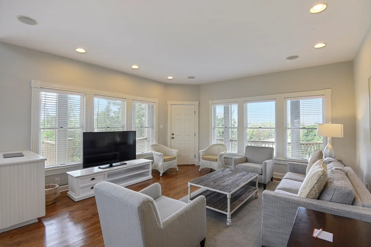 A spacious living room filled with natural light, featuring a comfortable seating arrangement centered around a coffee table, a television, and large windows that let the sun stream in. The inviting atmosphere suggests scenes of relaxation, group conversations, and cozy movie nights.