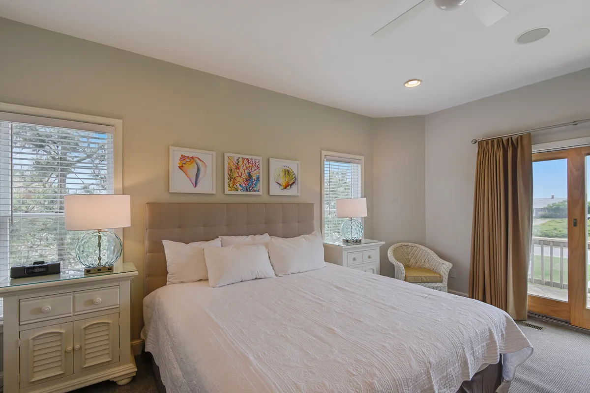A tranquil bedroom with a king-size bed dressed in soft linens, accented by tasteful artwork. Large windows allow natural light to fill the space, and the cozy chair in the corner offers a relaxing nook for reading or unwinding. A bedside lamp and subtle decor create a restful atmosphere.